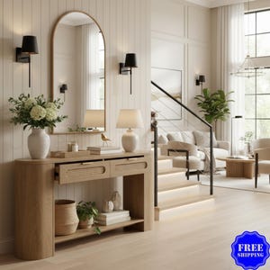 May include: A light oak console table with two drawers featuring woven cane panels and gold hardware. The table has a lower shelf holding a woven basket, a potted plant, and books. An arched mirror and two black wall sconces are mounted above.