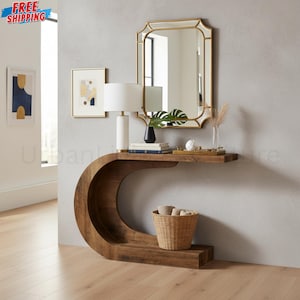 May include: A C-shaped wooden console table with a smooth finish, featuring a flat top surface and a curved base. A white cylindrical lamp, a stack of books, a small black vase, and a decorative tray are placed on the table surface.