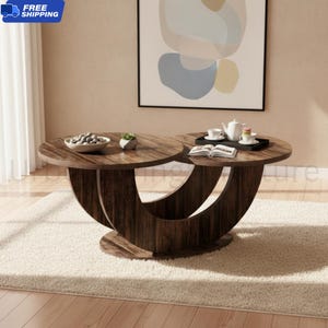 May include: A dark wood coffee table featuring two circular tabletops supported by a curved, crescent-shaped base. The table holds a white bowl filled with grey stones, a small succulent, a white teapot, two white cups, and an open book.