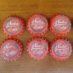 Op de afbeelding: Tien ronde metalen kroonkurken gerangschikt in twee rijen van vijf. Elke dop is rood geverfd met een geschulpte rand en is voorzien van de witte tekst Nuka Cola in een gestileerd lettertype op het midden van de bovenzijde van de dop.