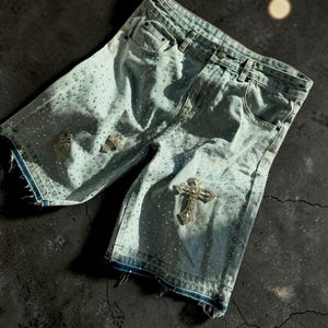 May include: Light blue denim shorts with a distressed look, featuring a cross-shaped embellishment and scattered rhinestones. The shorts have a faded wash and frayed edges, creating a unique, edgy style.