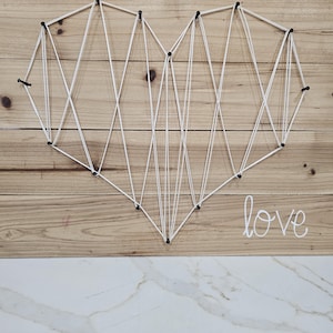 May include: A string art heart design created with white thread wrapped around black nails on a light brown wooden board. The word love is written in white cursive script in the lower right corner of the wooden surface.