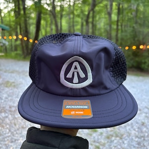 Può includere: Cappellino da baseball blu navy con logo bianco ricamato e retro in rete nera. Il cappellino ha una visiera curva e un'etichetta Richardson con le parole "B-Active" e "Wicking".