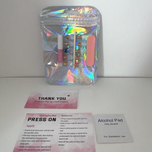 May include: A holographic zip bag containing a press-on nail kit. The kit includes nail glue, a nail file, and a set of light blue and gold press-on nails. Also included are instruction cards and an alcohol pad.
