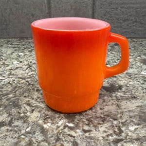 May include: An orange glass mug with a handle. The mug has a gradient color, transitioning from a lighter pink inside to a vibrant orange on the exterior. The mug is approximately 4 inches tall.