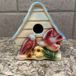 May include: A ceramic birdhouse figurine featuring a light blue roof and cream-colored walls. Two pink birds are sculpted on the front, one perched and one in flight, positioned near a central circular opening and a yellow flower with green leaves.