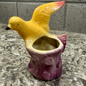 May include: A ceramic planter shaped like a yellow bird perched on a purple tree stump. The bird has a black beak and a wing extended upward with a pinkish-orange tip. The hollow opening for a plant is located in the center of the tree stump base.