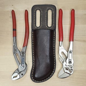 May include: A dark brown leather tool sheath with two belt slots at the top, positioned between two sets of metal pliers. Each pair of pliers features silver-coloured metal jaws and bright red rubberised handles for a secure grip during use.