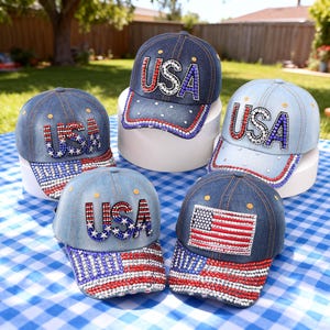Puede incluir: Cinco gorras de béisbol de mezclilla con adornos de pedrería. Cuatro gorras muestran el texto USA en pedrería, mientras que una presenta una bandera estadounidense de pedrería. Las viseras están decoradas con patrones de pedrería roja, blanca y azul.