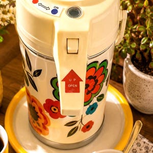 Könnte beinhalten: Eine cremefarbene elektrische Thermokanne mit einem lebendigen Blumenmuster in Rot, Orange und Blau. Der Deckel verfügt über einen OFF und ON Schalter mit einem roten und blauen Punkt sowie einen Hebel mit einem roten Pfeil und der Aufschrift UP OPEN.