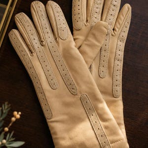 May include: A pair of light tan gloves featuring perforated leather detailing along the fingers and the back of the hand. The gloves have a smooth, matte finish and a structured design with stitched seams running vertically down each finger.