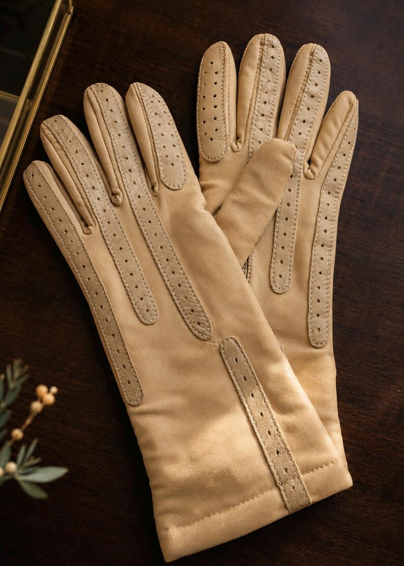 May include: A pair of light tan gloves featuring perforated leather detailing along the fingers and the back of the hand. The gloves have a smooth, matte finish and a structured design with stitched seams running vertically down each finger.