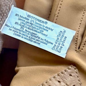 May include: Tan Isotoner gloves featuring a light brown knit cuff and a smooth exterior. A white care label is attached, displaying the brand name, material composition of 80% nylon and 20% spandex, lining details of 80% acrylic and 20% nylon, and one size.