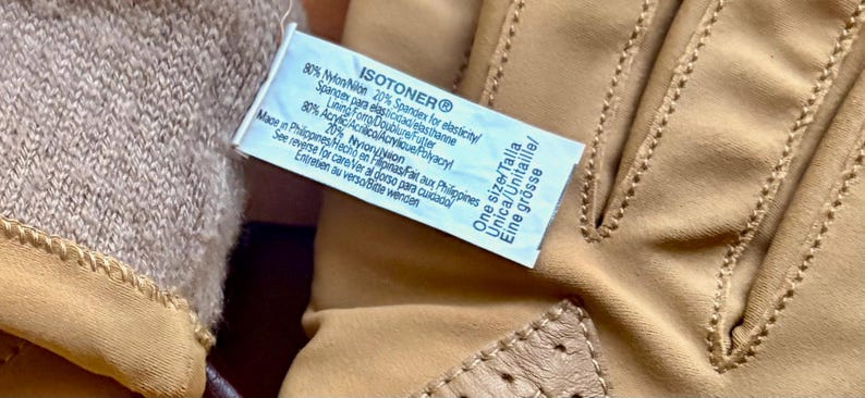 May include: Tan Isotoner gloves featuring a light brown knit cuff and a smooth exterior. A white care label is attached, displaying the brand name, material composition of 80% nylon and 20% spandex, lining details of 80% acrylic and 20% nylon, and one size.