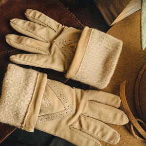 May include: A pair of beige leather gloves featuring a soft, textured knit cuff and decorative perforated detailing on the back of the hand. The gloves have a smooth finish and are designed with a classic construction, showcasing visible stitching.