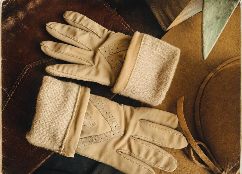 May include: A pair of beige leather gloves featuring a soft, textured knit cuff and decorative perforated detailing on the back of the hand. The gloves have a smooth finish and are designed with a classic construction, showcasing visible stitching.
