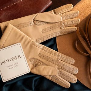 May include: A pair of tan gloves with perforated detailing on the fingers and palms. A white rectangular tag is attached to the gloves, featuring the text ISOTONER Famous for fit Since 1910 in black lettering, positioned on a dark fabric surface.