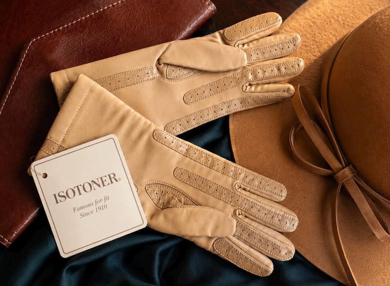 May include: A pair of tan gloves with perforated detailing on the fingers and palms. A white rectangular tag is attached to the gloves, featuring the text ISOTONER Famous for fit Since 1910 in black lettering, positioned on a dark fabric surface.