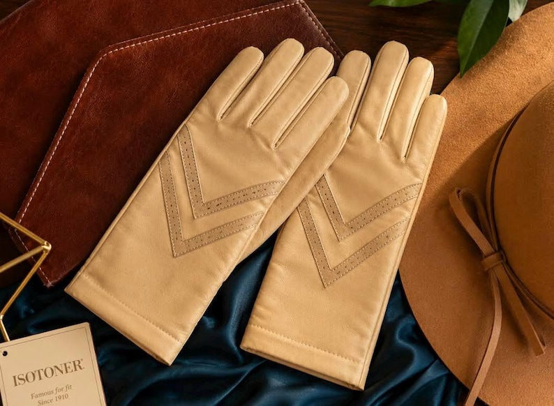 May include: A pair of beige leather gloves featuring a double chevron pattern with perforated detailing on the back of the hand. The gloves are accompanied by a rectangular tag with the text ISOTONER, Famous for fit, Since 1910 printed on it.