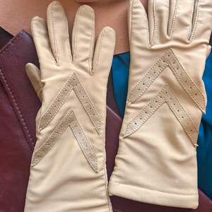 May include: A pair of beige gloves featuring a chevron-shaped cutout detail on the back of each hand. The gloves are constructed with smooth fabric and include stitched seams along the fingers and a straight hem at the wrist opening.