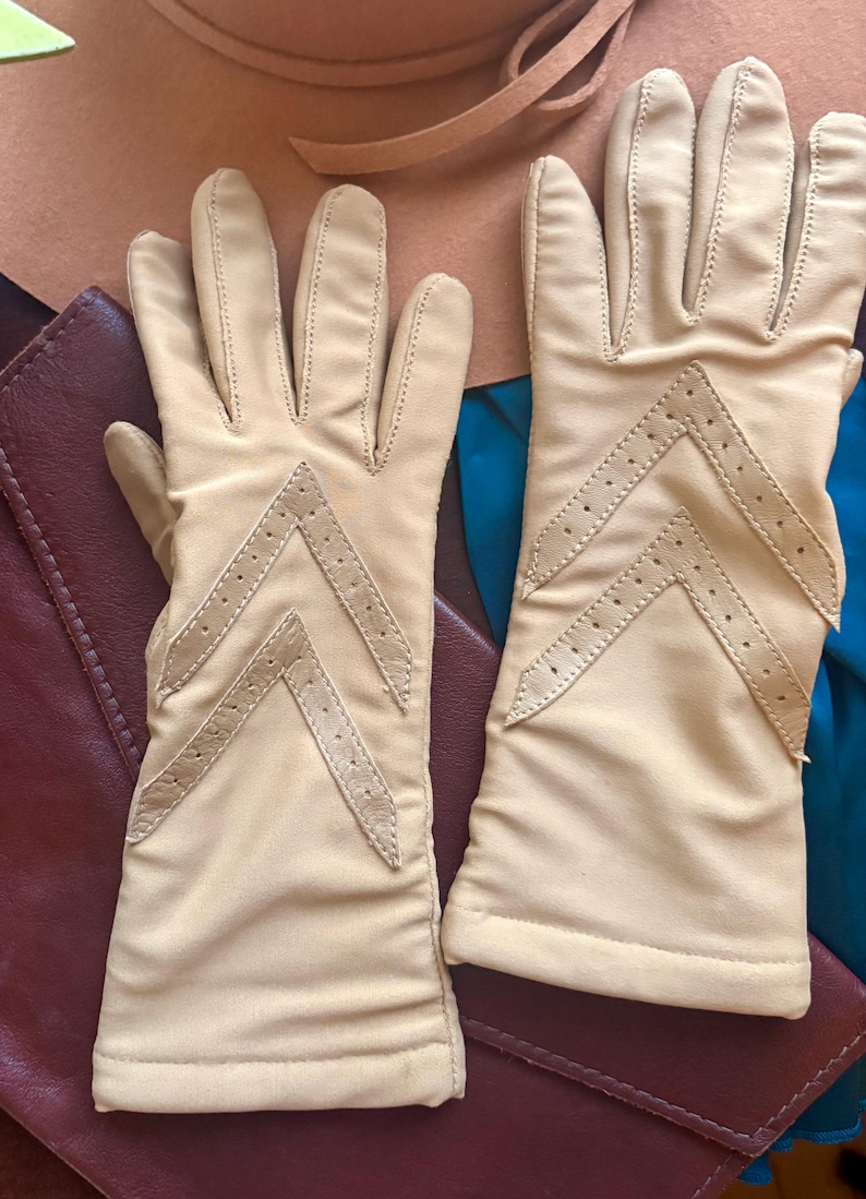 May include: A pair of beige gloves featuring a chevron-shaped cutout detail on the back of each hand. The gloves are constructed with smooth fabric and include stitched seams along the fingers and a straight hem at the wrist opening.