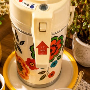 Può includere: Un distributore termico di bevande color crema con un motivo floreale in rosso, arancione, verde e blu. La parte superiore presenta un interruttore OFF/ON con punti rosso e blu e una leva con una freccia rossa e la scritta UP OPEN.