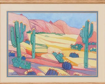 May include: A framed watercolor painting depicting a desert landscape with tall teal saguaro cacti and smaller round cacti with yellow flowers. The scene features rolling pink and tan hills under a light blue sky, all enclosed in a light wood frame.