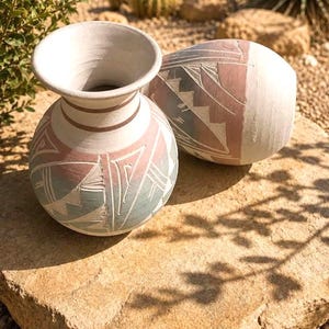 May include: Two ceramic vases featuring a matte white finish with etched geometric patterns in muted pink and sage green. The vases have a rounded body and a wide, flared rim, showcasing a hand-carved texture that reveals the clay underneath.
