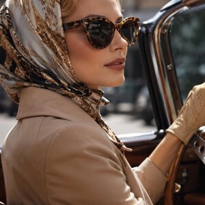 May include: A person wearing a beige coat, tan perforated leather gloves, and a silk headscarf featuring a white, black, and gold abstract pattern. The person is also wearing dark-tinted tortoiseshell sunglasses while sitting in a vehicle.