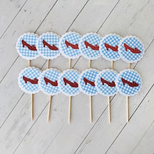 May include: Twelve red glitter ruby slipper cupcake toppers with blue and white gingham pattern on a white wood background.