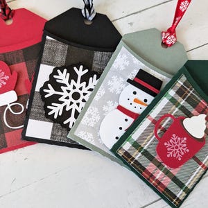 Puede incluir: Cuatro etiquetas de regalo navide&ntilde;as con elementos decorativos. Las etiquetas presentan un guante rojo, un copo de nieve blanco, un mu&ntilde;eco de nieve y una taza de chocolate caliente. Cada etiqueta tiene un patr&oacute;n diferente de cuadros o copos de nieve y est&aacute; adornada con una cinta.