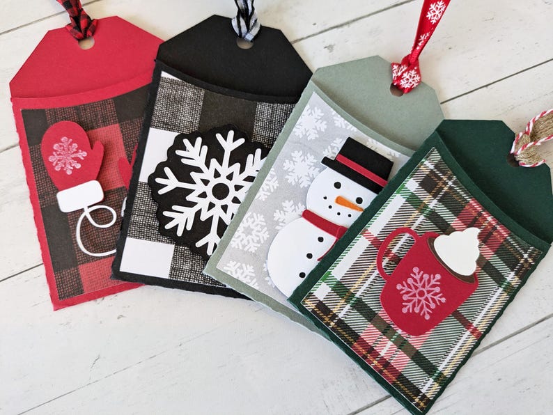 Puede incluir: Cuatro etiquetas de regalo navide&ntilde;as con elementos decorativos. Las etiquetas presentan un guante rojo, un copo de nieve blanco, un mu&ntilde;eco de nieve y una taza de chocolate caliente. Cada etiqueta tiene un patr&oacute;n diferente de cuadros o copos de nieve y est&aacute; adornada con una cinta.