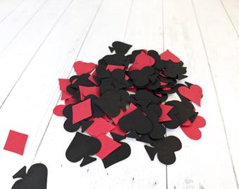 May include: A pile of black and red paper card suit cutouts, including hearts, spades, diamonds, and clubs. The cutouts are scattered on a white wooden surface.