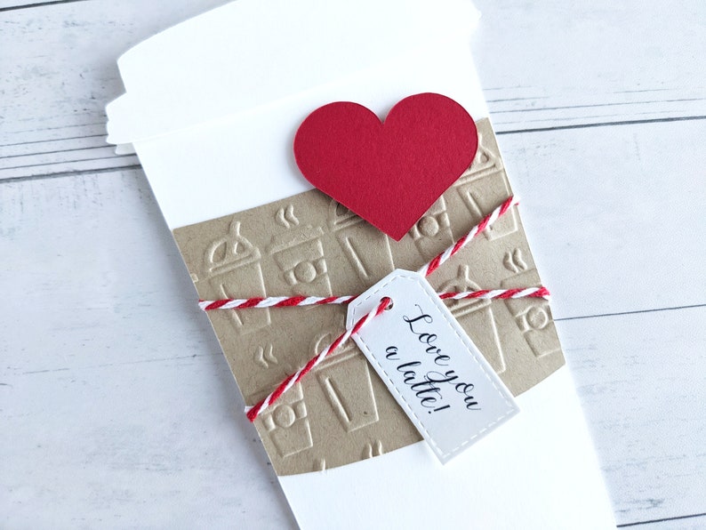 Puede incluir: Un vaso de caf&eacute; de papel blanco con una manga de papel marr&oacute;n decorada con iconos de vasos de caf&eacute;. La manga est&aacute; atada con una cuerda roja y blanca y una etiqueta blanca que dice "Love you a latte!" Un coraz&oacute;n rojo est&aacute; unido a la parte superior del vaso.