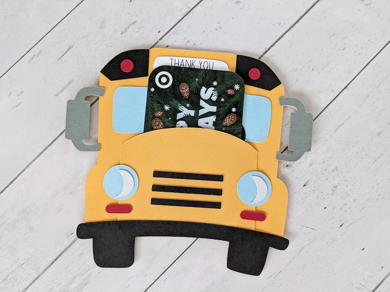 May include: A yellow school bus-shaped gift card holder with black wheels, trim, and accents. The bus has blue windows, headlights, and a red stop light. A gift card with a holiday design is visible inside the holder. The words "THANK YOU" are visible.