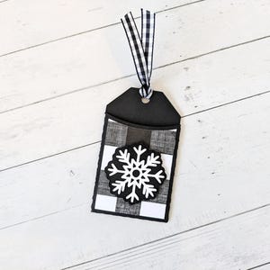 Puede incluir: Una etiqueta de regalo en blanco y negro con un dise&ntilde;o de copo de nieve. La etiqueta presenta un patr&oacute;n de cuadros de b&uacute;falo en blanco y negro y un copo de nieve blanco. Una cinta de cuadros vichy en blanco y negro est&aacute; unida a la parte superior.