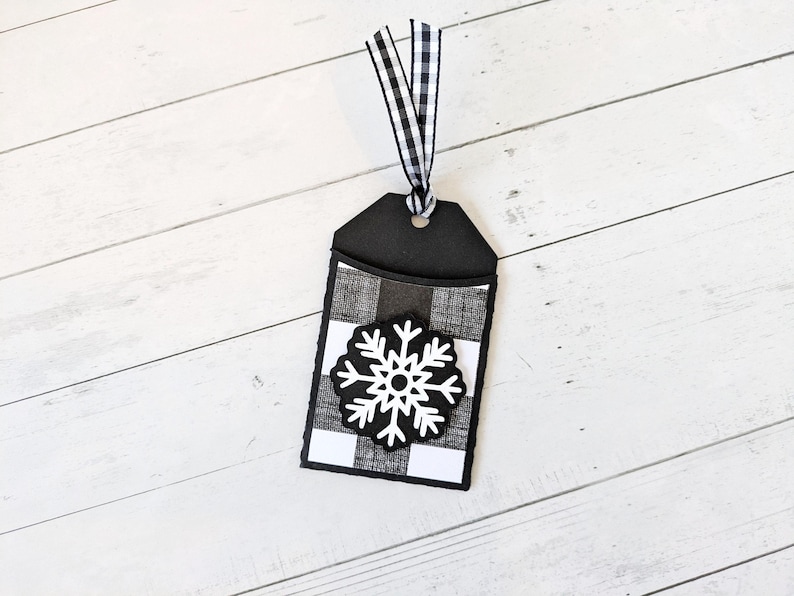 Puede incluir: Una etiqueta de regalo en blanco y negro con un dise&ntilde;o de copo de nieve. La etiqueta presenta un patr&oacute;n de cuadros de b&uacute;falo en blanco y negro y un copo de nieve blanco. Una cinta de cuadros vichy en blanco y negro est&aacute; unida a la parte superior.