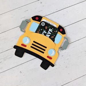 May include: A yellow school bus-shaped gift card holder with black wheels and accents. The bus has blue windows and a gift card with the words "Thank You" visible. The holder is on a white wooden surface.