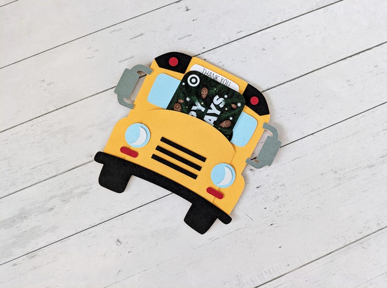 May include: A yellow school bus-shaped gift card holder with black wheels and accents. The bus has blue windows and a gift card with the words "Thank You" visible. The holder is on a white wooden surface.