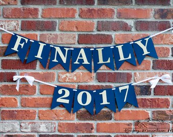 May include: A navy blue and gold graduation banner with the words "FINALLY 2017" hanging on a brick wall.
