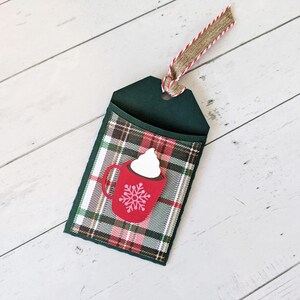 Puede incluir: Una etiqueta de regalo verde oscuro con un bolsillo a cuadros con un gr&aacute;fico de taza roja con crema batida y un copo de nieve. La etiqueta tiene una cinta de arpillera y roja para colgar. El fondo es una superficie de madera blanca.