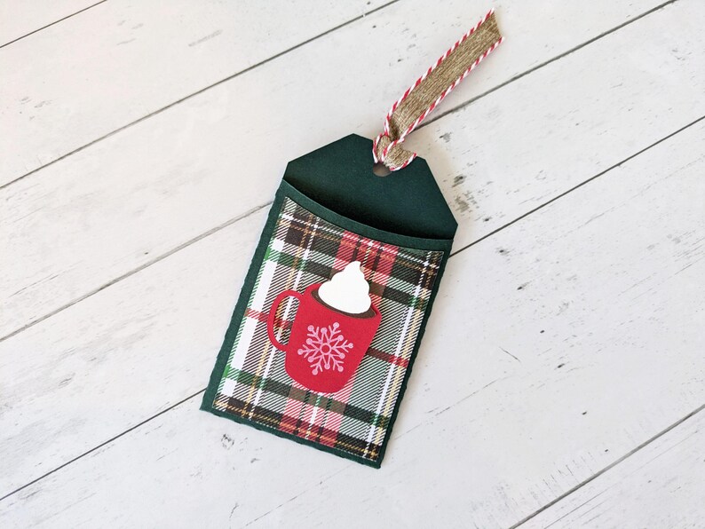 Puede incluir: Una etiqueta de regalo verde oscuro con un bolsillo a cuadros con un gr&aacute;fico de taza roja con crema batida y un copo de nieve. La etiqueta tiene una cinta de arpillera y roja para colgar. El fondo es una superficie de madera blanca.