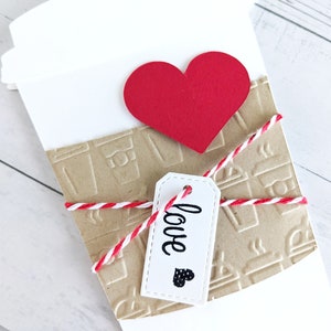 Puede incluir: Un vaso de papel blanco con una manga de papel marr&oacute;n decorada con dise&ntilde;os de vasos de caf&eacute;. El vaso est&aacute; atado con una cuerda roja y blanca y una etiqueta blanca que dice "love" con un peque&ntilde;o coraz&oacute;n negro. Un coraz&oacute;n de papel rojo se coloca en la parte superior del vaso.