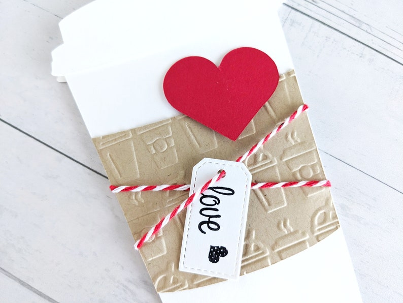 Puede incluir: Un vaso de papel blanco con una manga de papel marr&oacute;n decorada con dise&ntilde;os de vasos de caf&eacute;. El vaso est&aacute; atado con una cuerda roja y blanca y una etiqueta blanca que dice "love" con un peque&ntilde;o coraz&oacute;n negro. Un coraz&oacute;n de papel rojo se coloca en la parte superior del vaso.