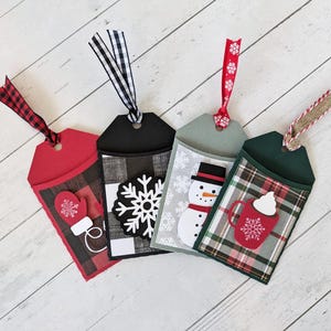 Puede incluir: Cuatro etiquetas de regalo navide&ntilde;as hechas a mano con bolsillos decorativos. Cada etiqueta presenta un dise&ntilde;o diferente: un guante rojo, un copo de nieve, un mu&ntilde;eco de nieve y una taza de chocolate caliente. Las etiquetas est&aacute;n hechas de cartulina y tienen detalles de papel estampado y cinta.