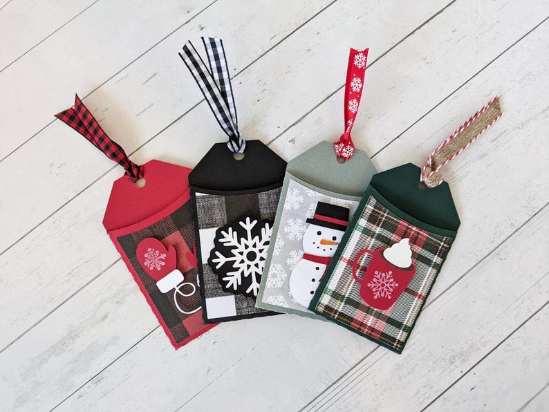 Puede incluir: Cuatro etiquetas de regalo navide&ntilde;as hechas a mano con bolsillos decorativos. Cada etiqueta presenta un dise&ntilde;o diferente: un guante rojo, un copo de nieve, un mu&ntilde;eco de nieve y una taza de chocolate caliente. Las etiquetas est&aacute;n hechas de cartulina y tienen detalles de papel estampado y cinta.