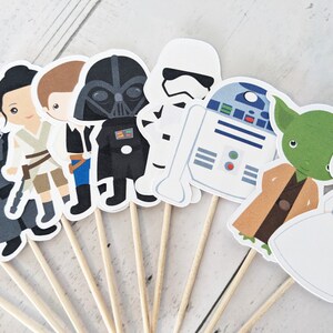 Puede incluir: Un conjunto de 12 adornos para cupcakes de personajes de Star Wars. Los adornos presentan personajes como Darth Vader, Yoda, R2-D2 y la princesa Leia. Los adornos est&aacute;n impresos en cartulina y unidos a brochetas de madera.