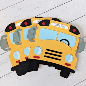 May include: Yellow school bus-shaped paper cutouts with black tyres and accents. Each cutout has blue windows, a red stop light, and the words "THANK YOU" on top. The cutouts are arranged on a white wooden surface.
