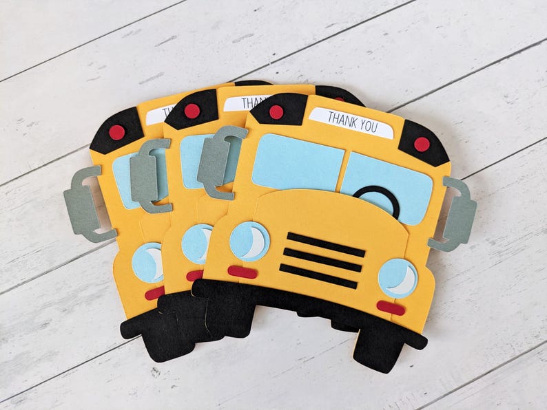 May include: Yellow school bus-shaped paper cutouts with black tyres and accents. Each cutout has blue windows, a red stop light, and the words "THANK YOU" on top. The cutouts are arranged on a white wooden surface.