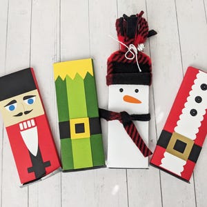 May include: Four Christmas-themed gift boxes. Designs include a nutcracker, an elf, a snowman, and Santa Claus. The boxes are red, green, white, and black with decorative elements like belts, hats, and facial features.